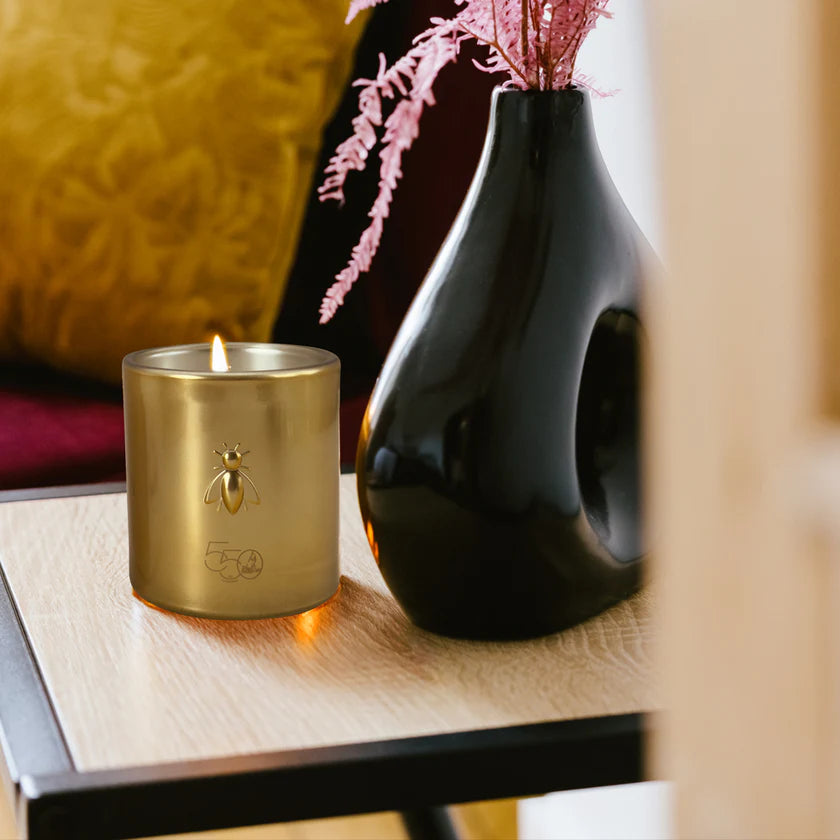 Gold candle with a bee logo on a wooden surface next to a black vase with pink flowers.